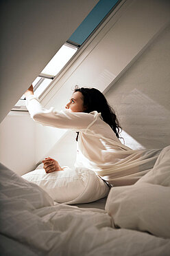 Frau zieht im Bett kniend das Plissee zum Sonnenschutz am Dachfenster herunter