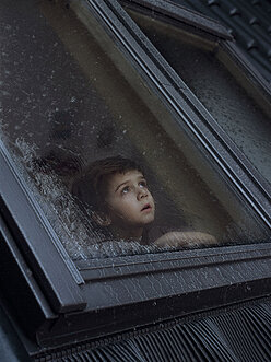 Kleiner Junge, der besorgt aus dem Dachfenster auf ein Unwetter schaut